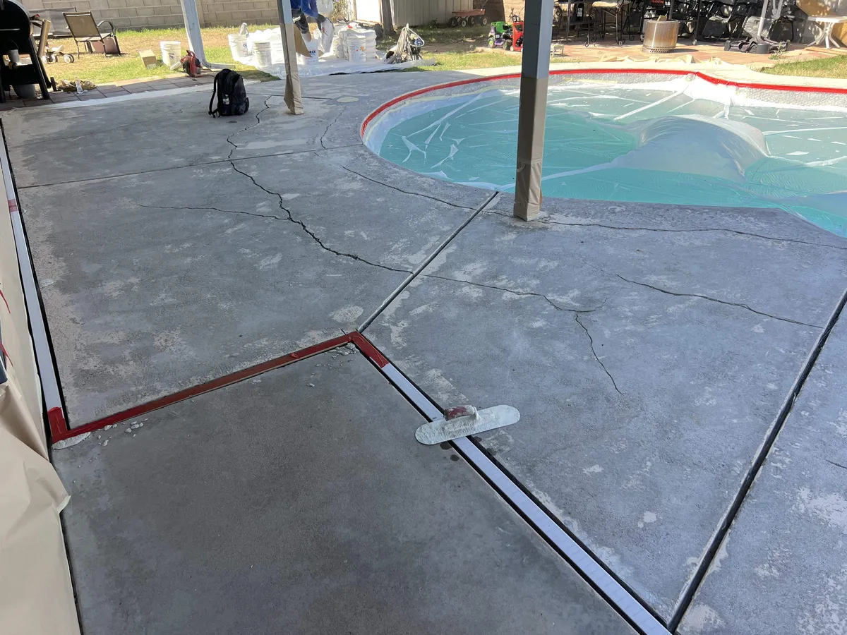 Cracked gray concrete pool deck in the Phoenix Valley before repair, with cracks wandering across the slab, the pool covered, and the trench drains taped off for prep